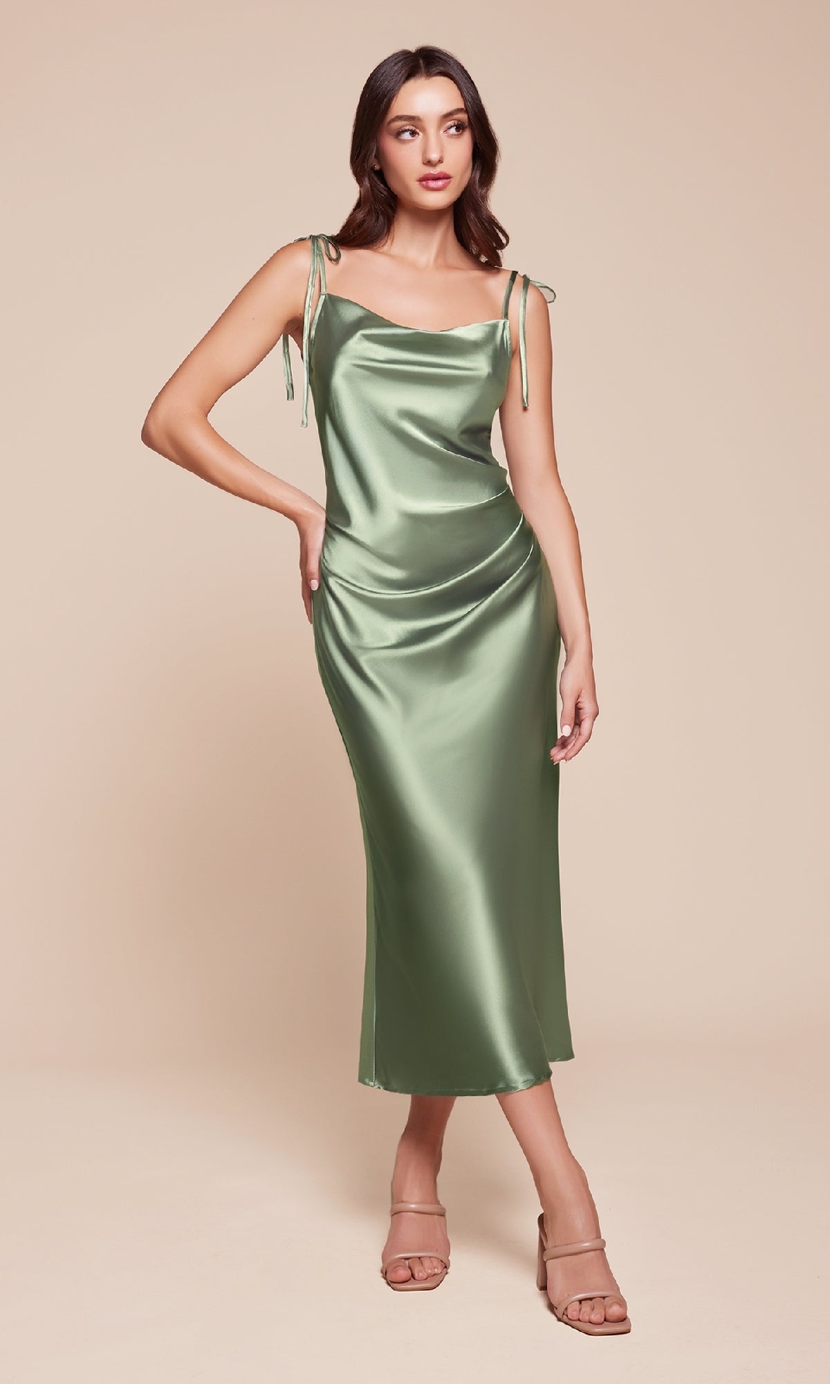 Woman wearing the Midi Wedding-Guest Slip Dress BD103 in light green satin with shoulder tie straps and beige heeled sandals, standing against a plain beige background.